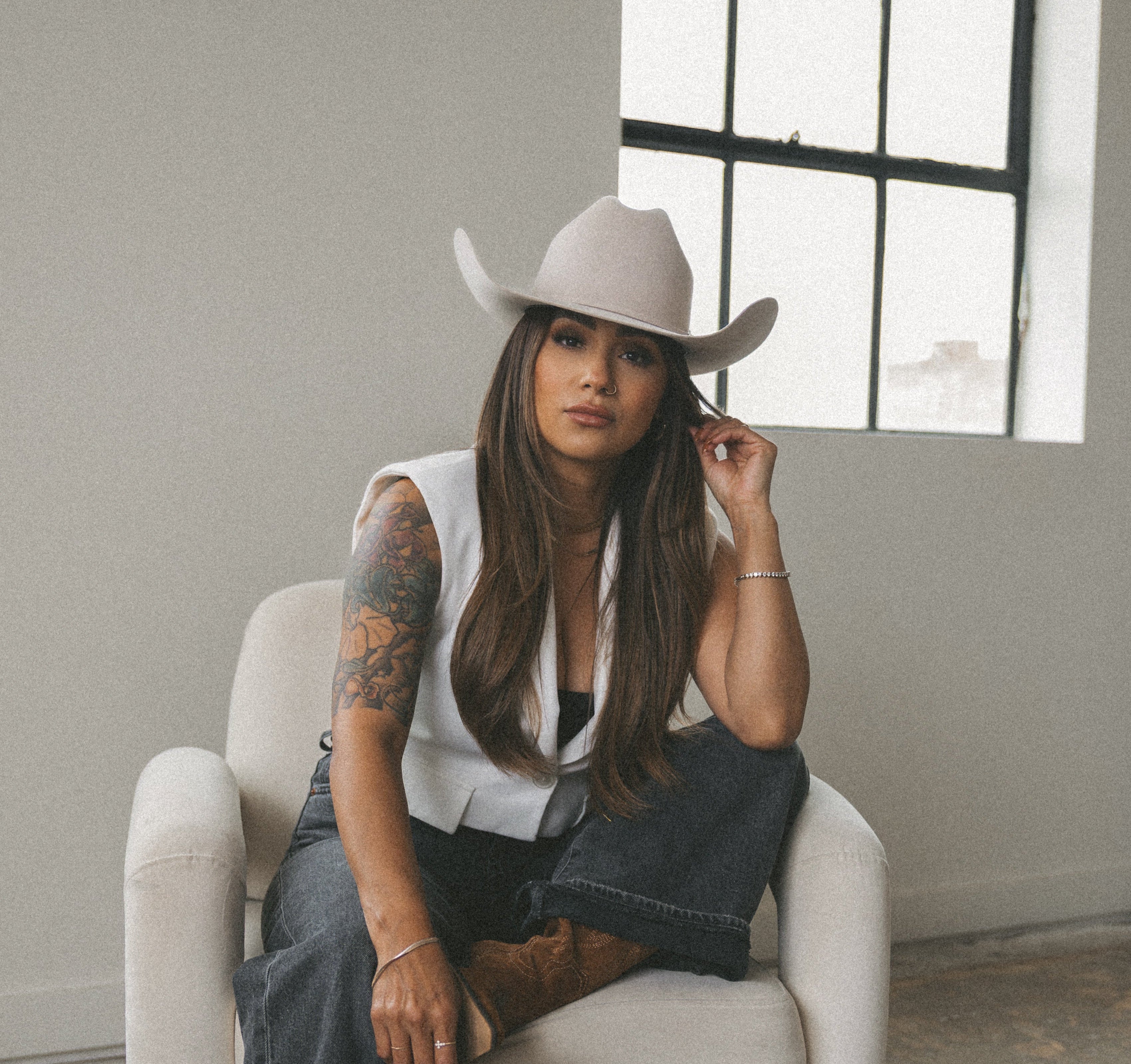 A woman wearing a light beige cowboy hat, a white sleeveless top, dark blue jeans, and brown boots sits in a white armchair in a minimalist room with a large window.